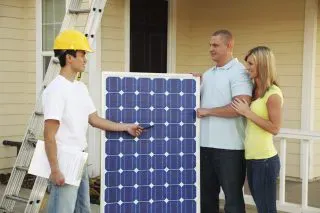 Ein Installateur zeigt einem Paar vor seinem Haus, das neben einer Leiter steht, ein Solarmodul und verdeutlicht, wie ein Online-Preisrechner für Solaranlagen die Planung erleichtern kann.